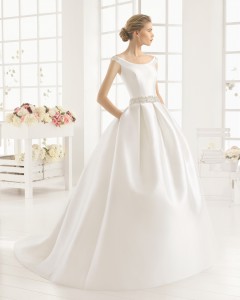 Brudekjole i kraftig brudekjole satin med bådudskæring, krystalbælte samt fyldigt skørt med lommer samt slæb.